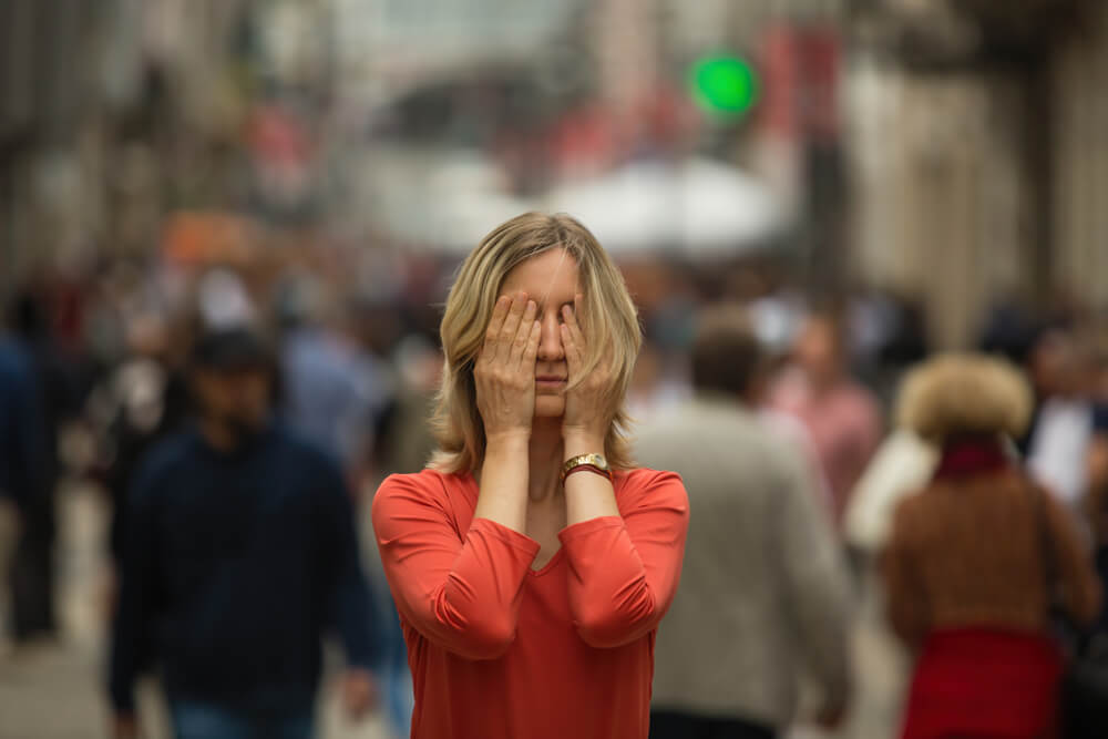 panic attack A woman in a red shirt covering her eyes, standing still in the middle of a busy sidewalk